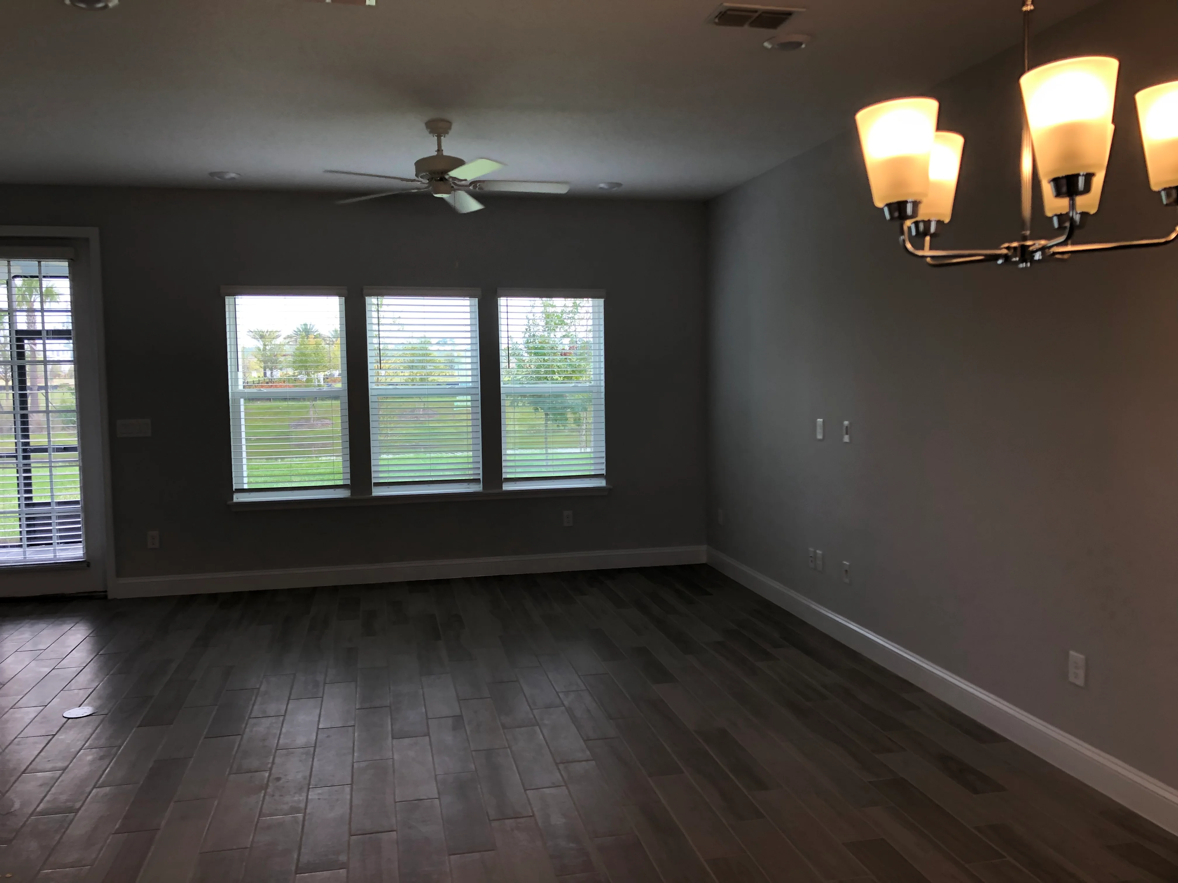 Spacious room with dark wood flooring, gray walls, modern lighting fixtures, and large windows with blinds