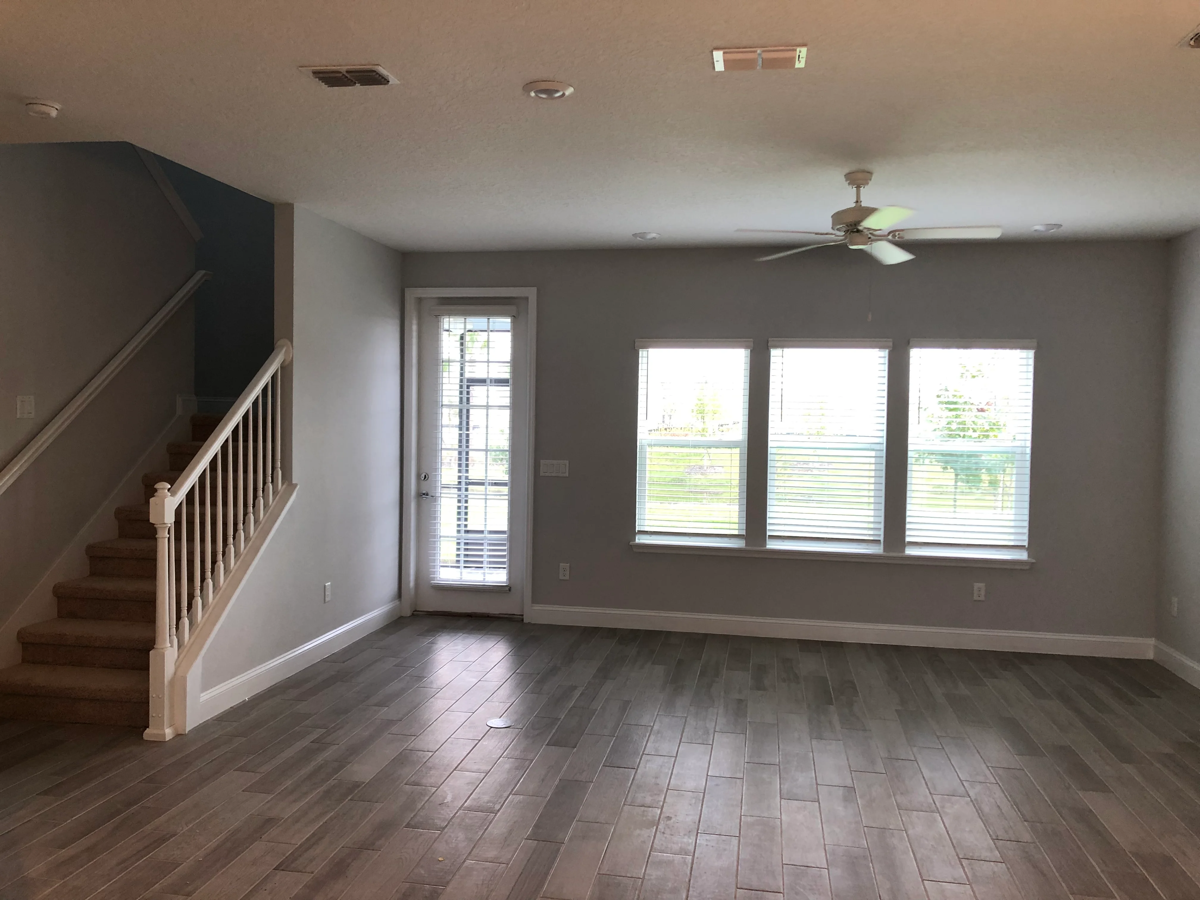 Open living room with herringbone tile flooring, staircase, and three large windows with shutters